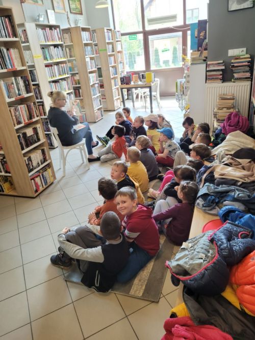 na zdjęciu widoczna grupa dzieci siedząca w bibliotece, naprzeciw nich koło regałów siedzi bibliotekarz