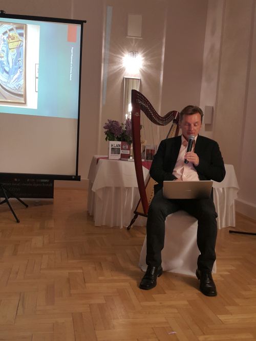 na zdjęciu widoczny mężczyzna siedzący na krześle, w ręce trzyma mikrofon, czyta z laptopa który leży na jego kolanach, w tle stoliki i wyświetlana prezentacja