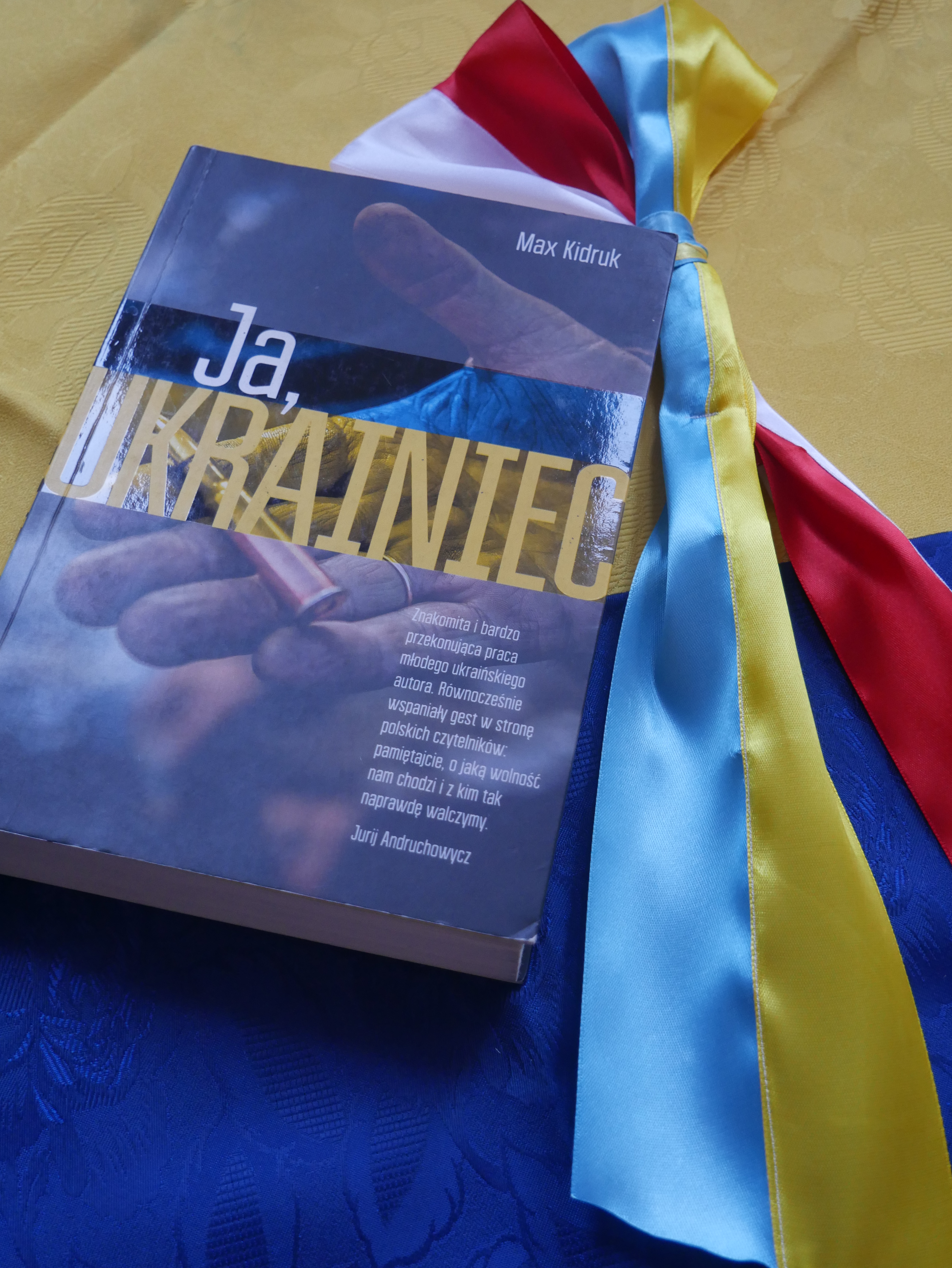 zdjęcie przedstawia książkę leżącą na żółto-niebieskim tle, obok książki leżą kokardy narodowe Polski i Ukrainy
