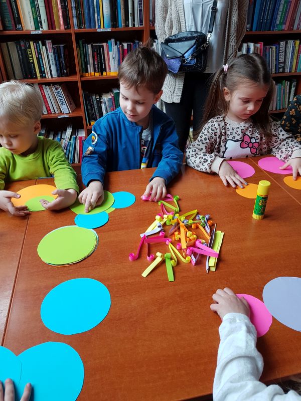 zdjęcie przedstawia dzieci siedzące przy stole podczas zajęć plastycznych w bibliotece