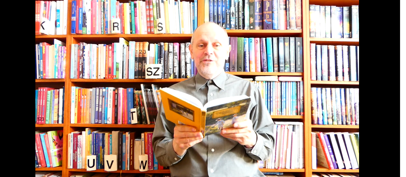 Mężczyzna w jasnej koszuli i Mężczyzna w szarej koszuli siedzi z otwartą książką na tle półek z kolorowymi książkami.