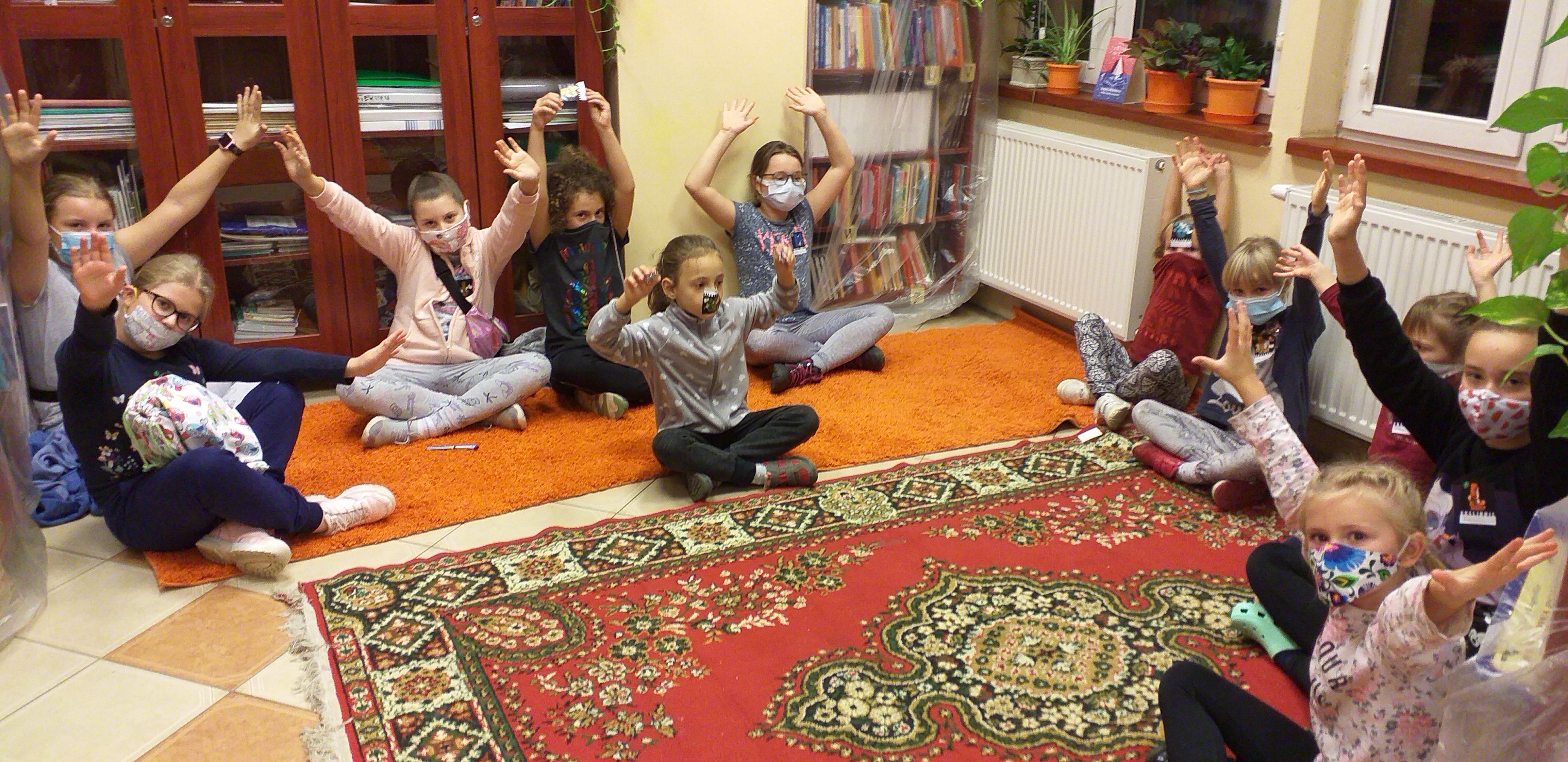Jedenaścioro dzieci siedzi po turecku na podłodze z uniesionymi rękami, część trzyma się za ręce. Na środku kolorowy dywan.