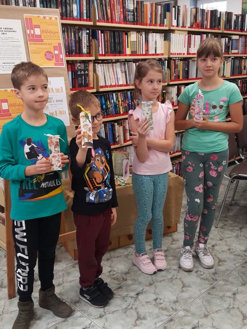 czwórka dzieci na tle regałów z książkami, pokazują prace wykonane na warsztatach