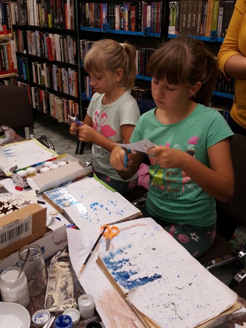 dwie dziewczynki wyknują prace plastyczne, w tle regały biblioteczne