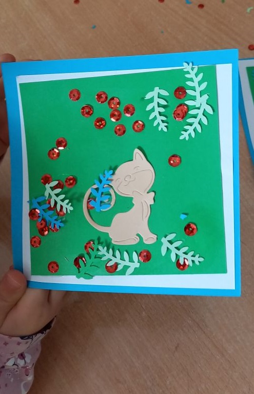 zdjecie przedstawia kartkę z kotem ozdocioną kolorowymi listkami i cekinami, kartkę trzyma w ręce dziecko nad stolikiem