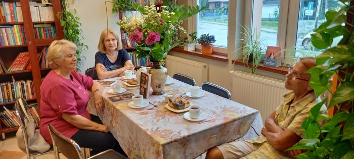 na zdjęciu widiczne trzy kobiety siedzące przy stole, na stole wazon z kwiatami, książki, filiżanki i tależyki