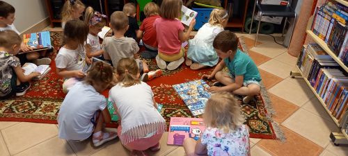 na zdjęciu widoczna grupa dzieci siedząca z ksiązkami na dywanie w bibliotece