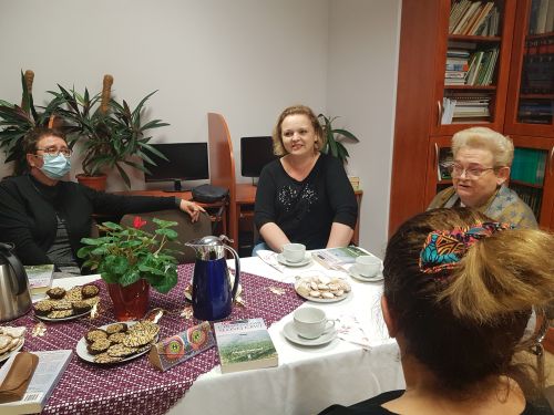 cztery kobiety siedzą przy stole, jedna jest odwrócona tyłem, na stole filiżanki z herbatą i ciasto, trwa dyskusja