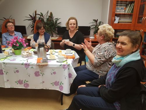 pięć kobiet w średnim wieku siedzi przy stole, ma stole przed nimi dzbanek, ciastka, książki i kwiaty