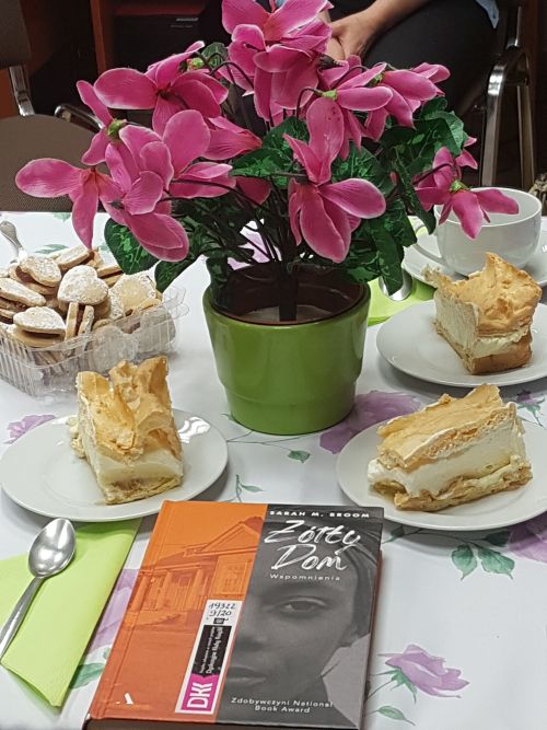 na stole leży książka, za nią różowe kwiaty, w koło ciasto na tależykach i ciasteczka