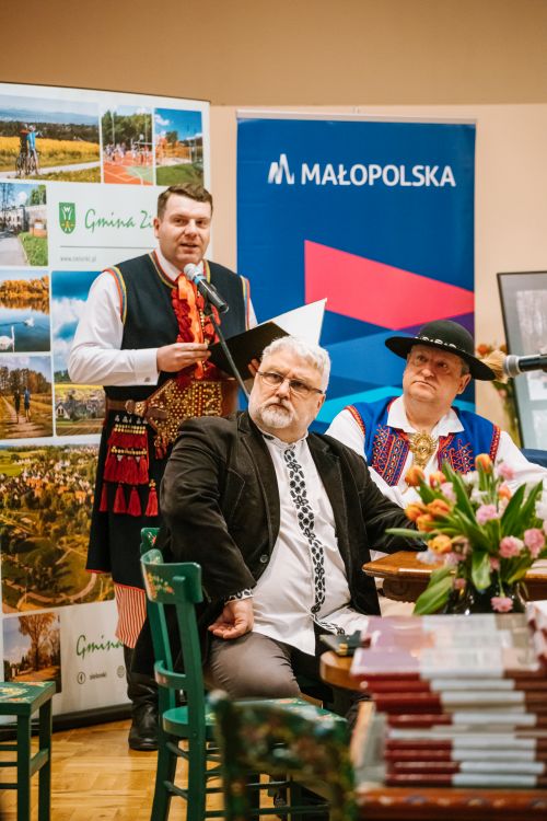 dwóch mężczyzn stoi przy stole, jeden z nich jest ubrany w strój ludowy, za nimi stoi mężczyna ubrany w strój krakowski, w tle banery z napisami Małopolska i Gmina Zielonki