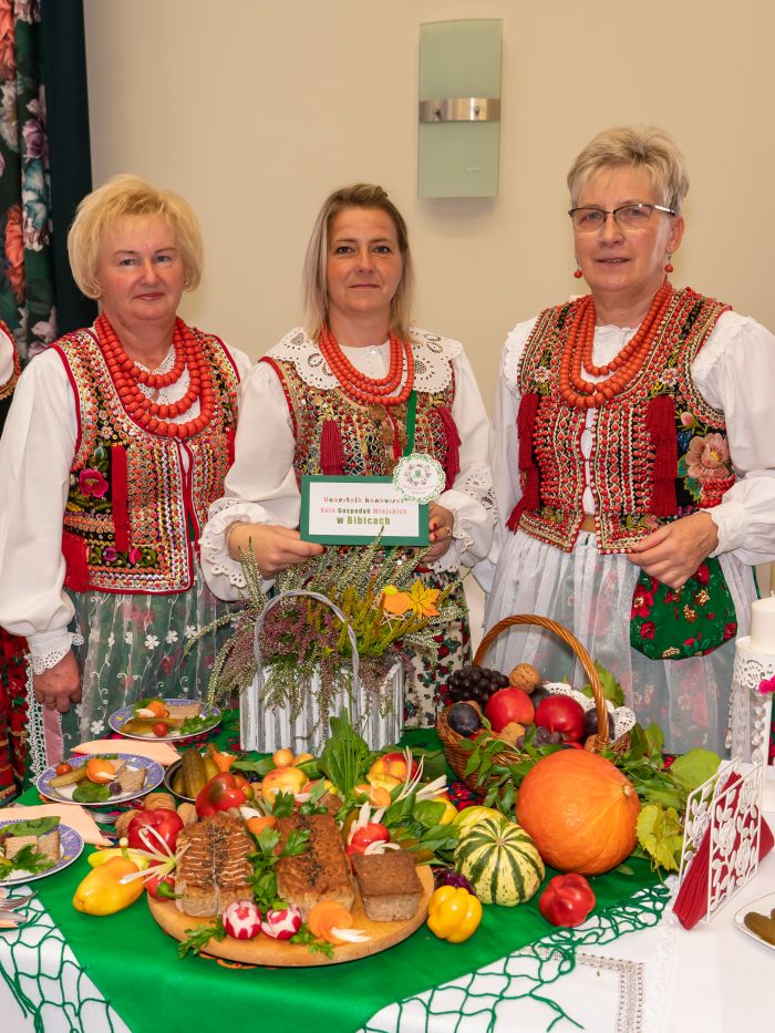 zdjęcie przedstawia trzy kobiety w strojach krakowskich stojące przed stolikiem z jedzeniem i dekoracjami
