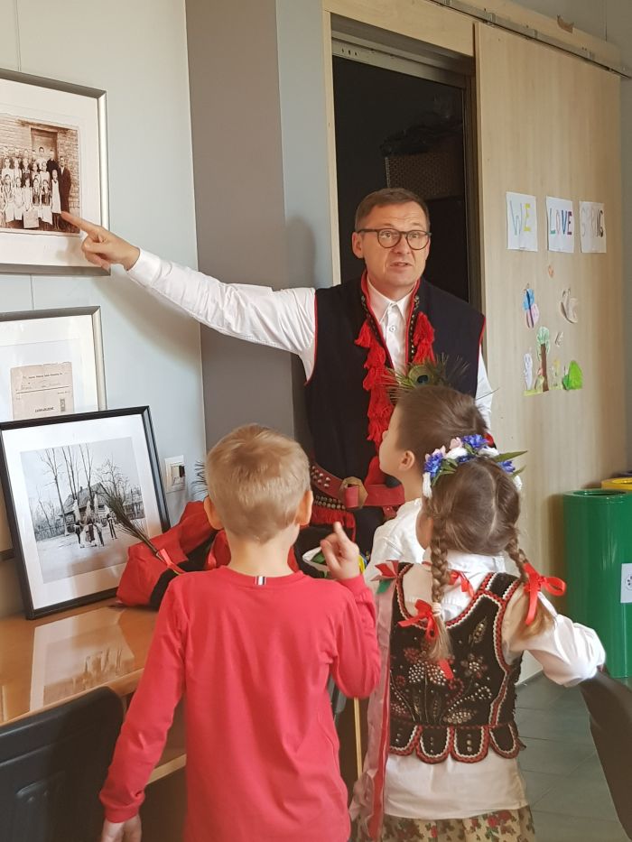 zdjęcie przedstawia mężczyznę w stroju krakowskim prezentującego dzieciom obrazy na ścianie