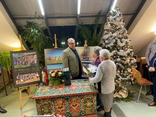 w tle wystawa fotografii i dekoracje świąteczne, dwie kobiety składają życzenia mężczyźnie, cała trójka stoi obok skrzyni krakowskiej