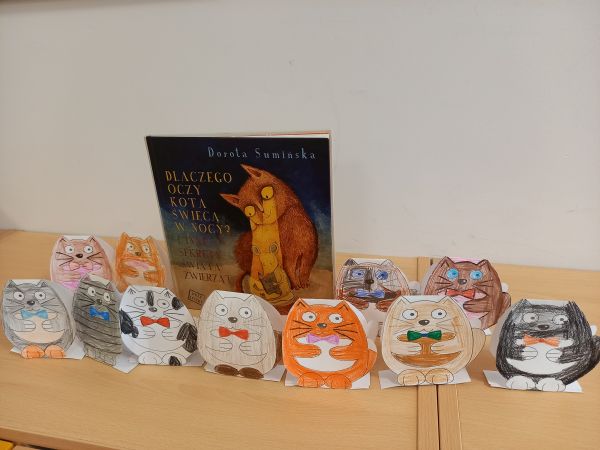 zdjęcie przedstawia książkę z kotem na okładce stojącą wśród kotów z papieru wykonanych przez dzieci
