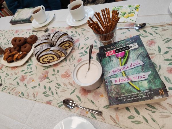 zdjęcie przedstawia stół, na nim książkę, filiżanki z herbatą i ciasto