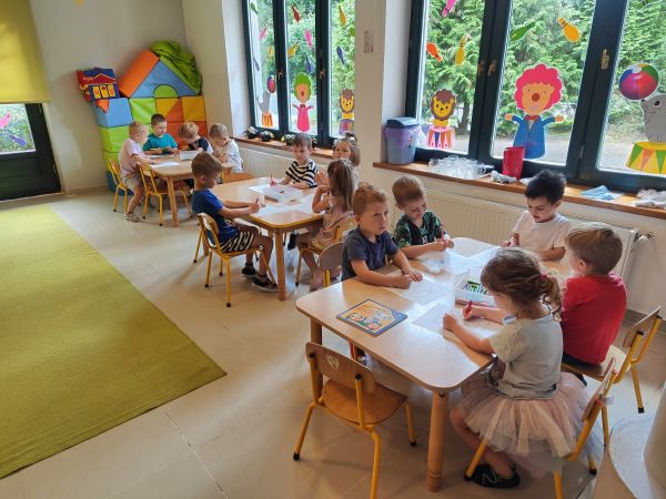 zdjęcie przedstawia grupę przedszkolaków siedzącą przy stolikach podczas zajęć w przedszkolu