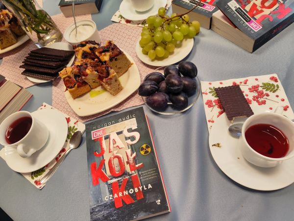 zdjecie przedstawia książkę leżącą na stole w otoczeniu ciasta i filiżanek z herbatą