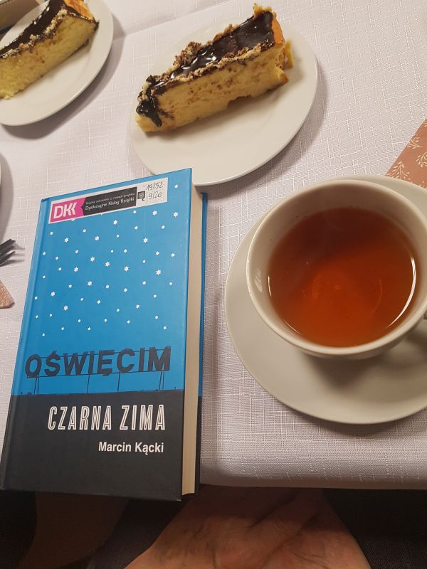 zdjęcie przedstawia książkę, filiżankę z herbatą i talerzyk z ciastem leżące na stole