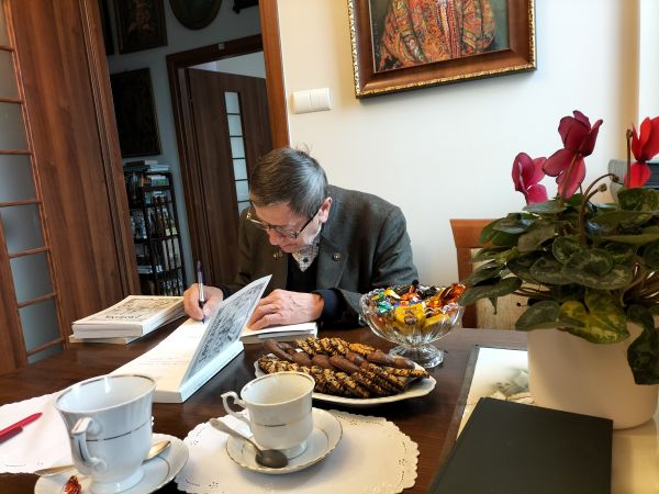 na zdjęciu widoczny starszy mężczyzna siedzący przy stoliku i podpisujący książkę