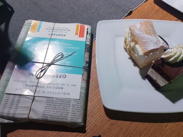 zdjęcie przedstawia książkę zapakowaną w gazetę z ulotką i talerzyk z ciastem