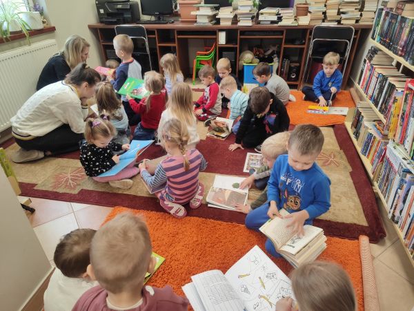 zdjęcie przedstawia przedszkolaki siedzące na dywanie w bibliotece i oglądające książki 
