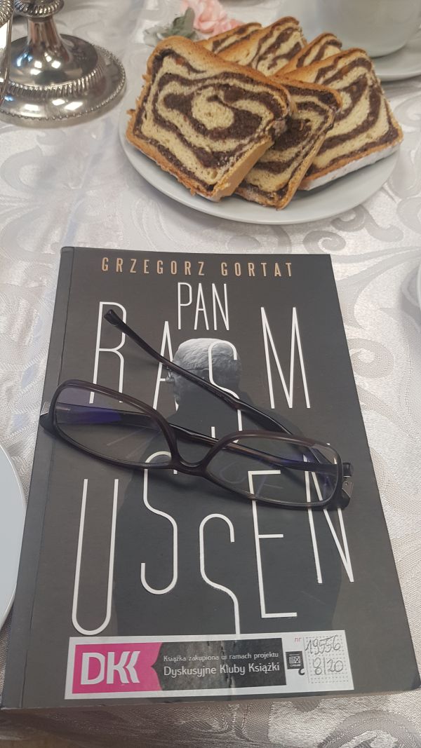 zdjęcie książki leżacej na stole, na niej leżą okulary, obok tależ z ciastem