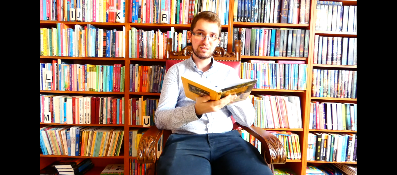 Mężczyzna w jasnej koszuli i granatowych spodniach siedzi z otwartą książką na tle półek z kolorowymi książkami.